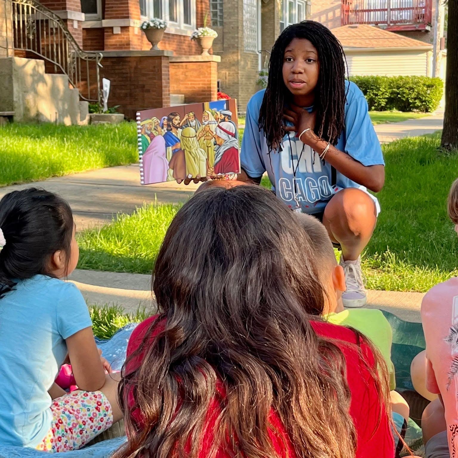A teenage leader crouches on a sidewalk and shows a picture book to children sitting on the grass