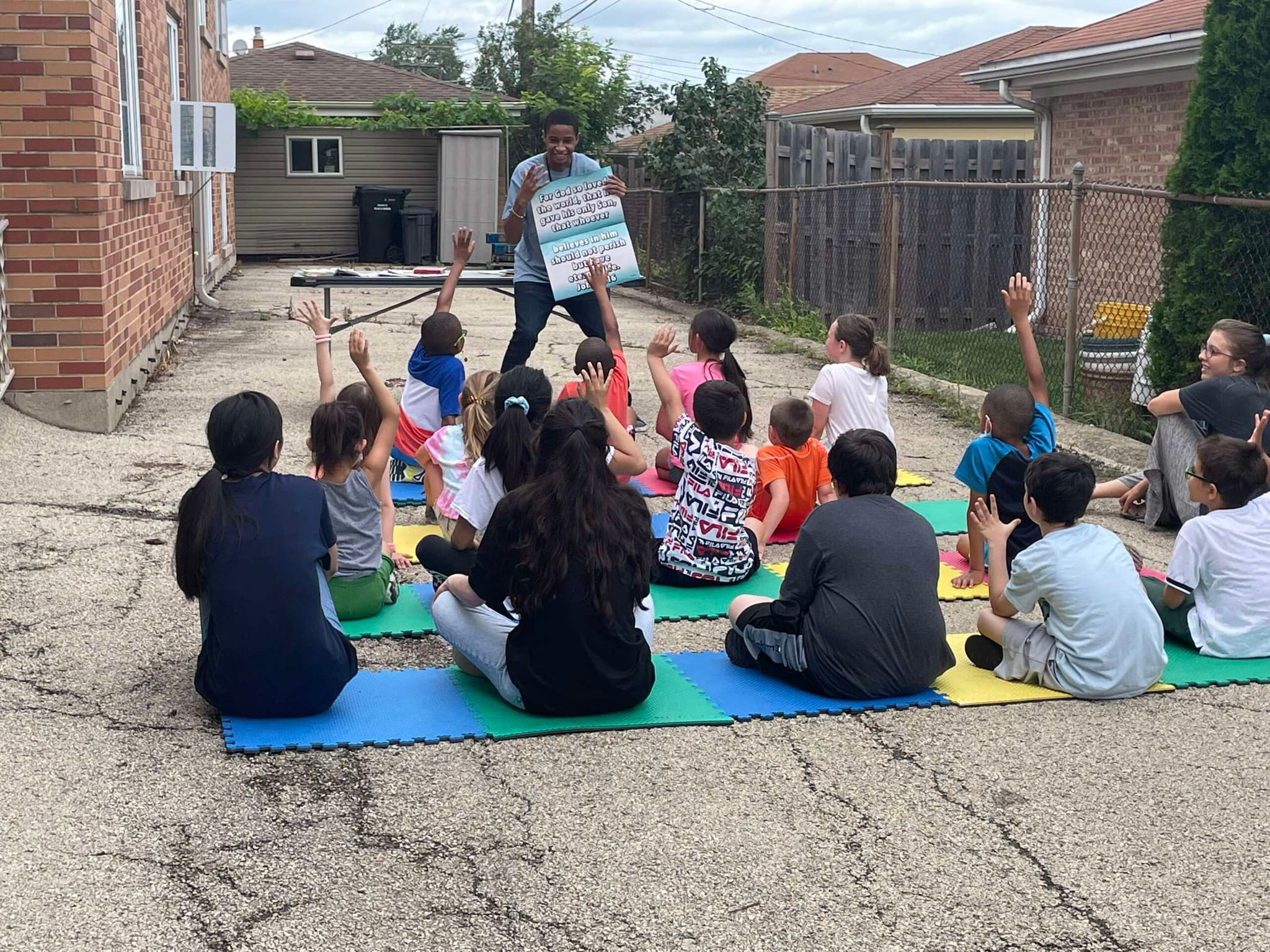 A teacher holds up a lesson card for children sitting on mats in a driveway, hands raised