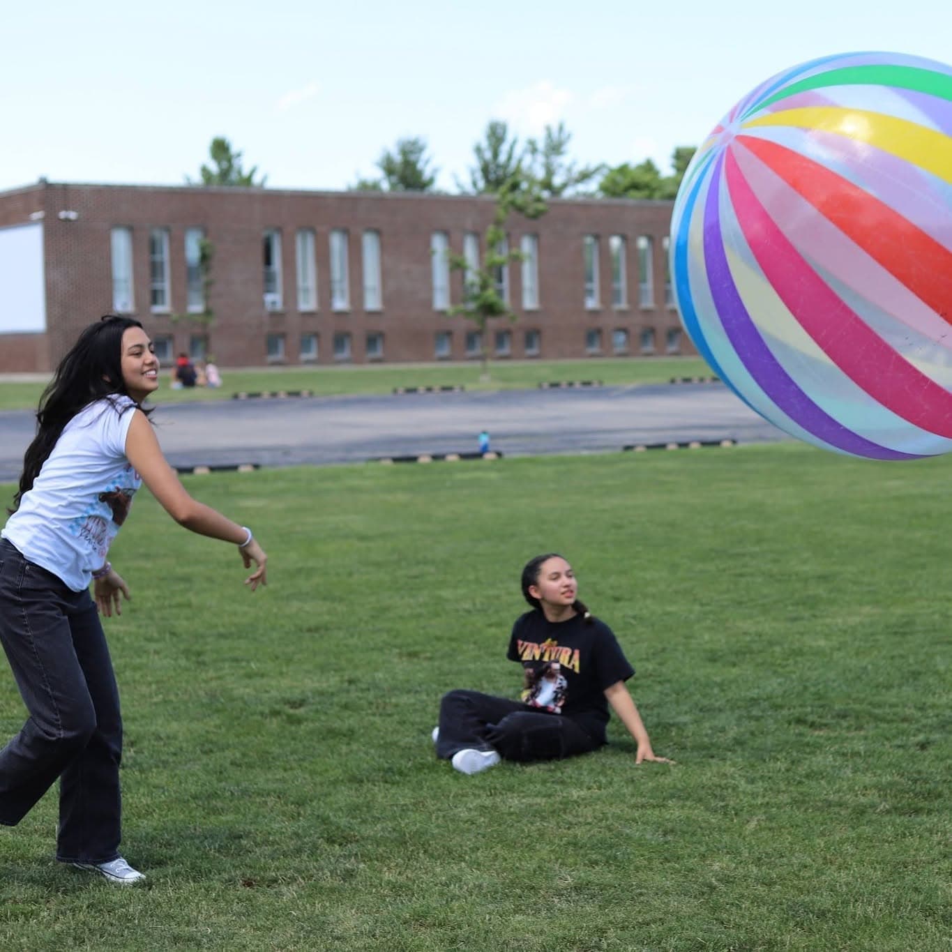 A girl throws a giant striped beach ball across a field while another sits on the grass