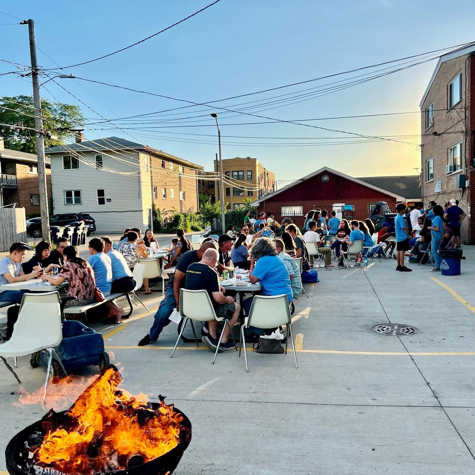 Neighbors sit at tables in a parking lot between apartment buildings at sunset, a fire pit in the foreground