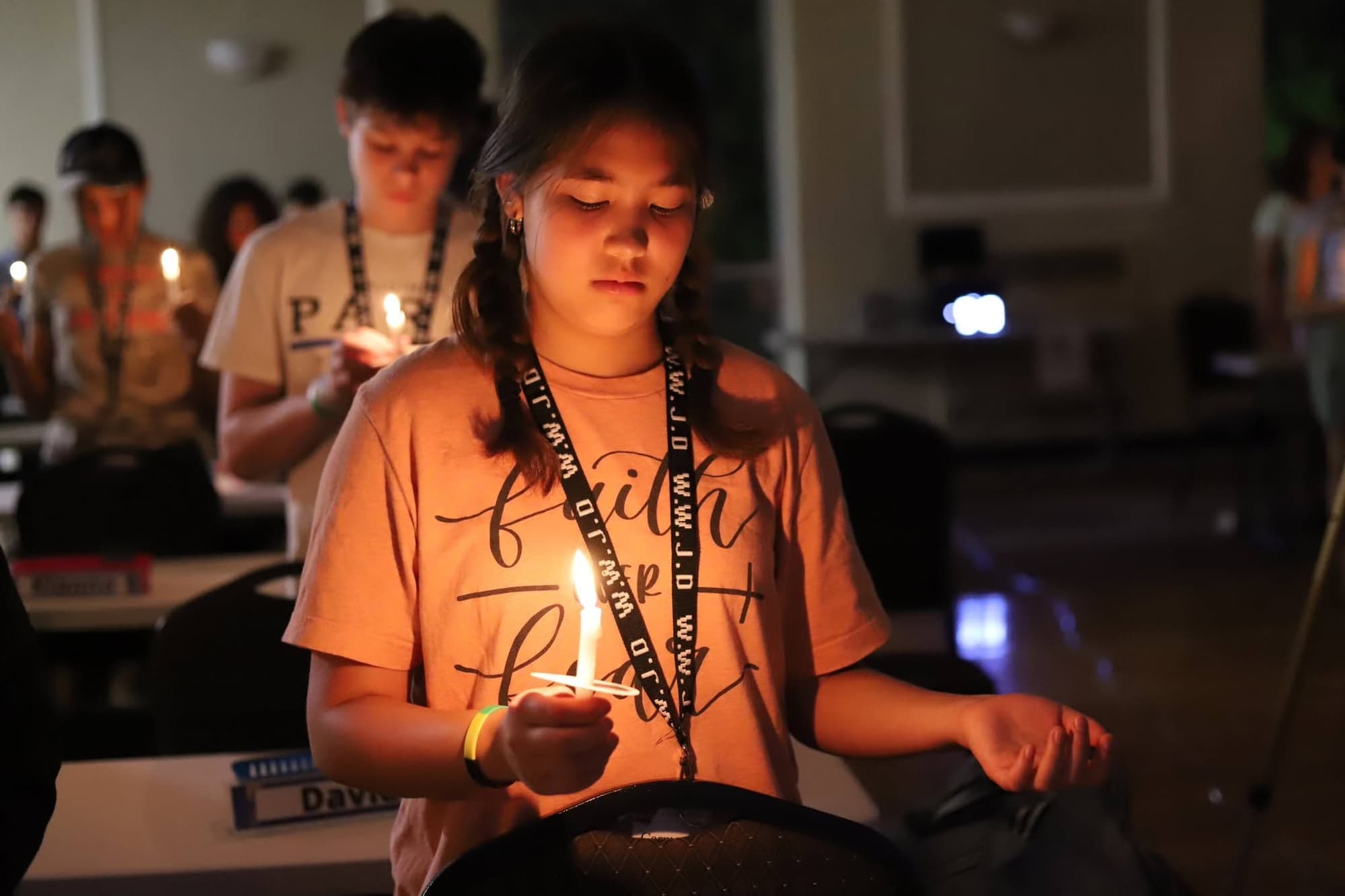 A teenager holds a lit candle in a darkened room, others with candles behind her