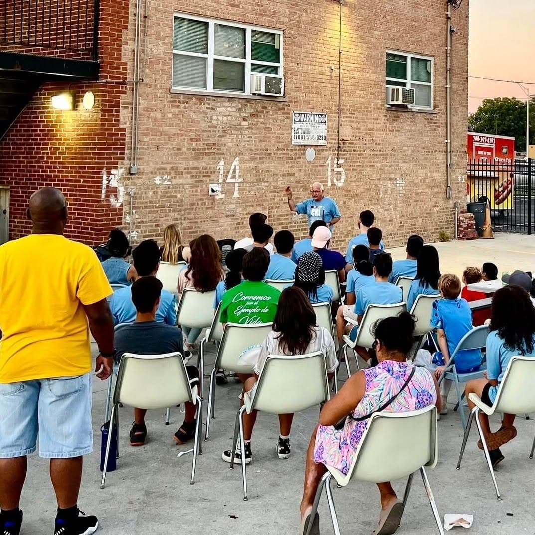 Families sit on chairs in a courtyard at dusk and listen to a speaker in front of a brick wall