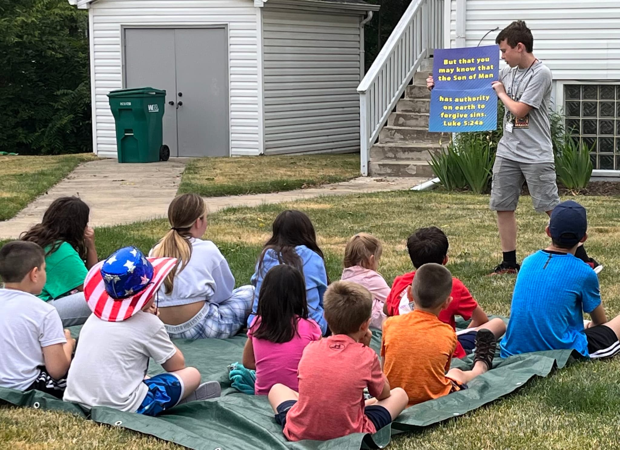 5-Day Club in a neighborhood: children sit on a tarp on the grass while a teacher holds up a verse